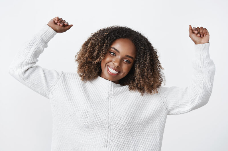 Do victory dance. Charming energized and active positive dark-skinned sportswoman raising hands up in joy and amusement smiling broadly dancing feeling upbeat wearing sweater over white wall.